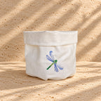 A small off-white fabric storage basket with a folded top edge and an embroidered dragonfly on the front, set on a sandy surface with dappled shadows. MI RE LA