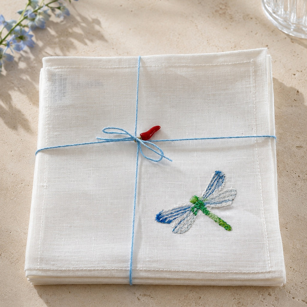 A folded white linen napkin with a blue dragonfly embroidery on the corner, tied with a blue string and a small red bow, set on a beige stone surface. MI RE LA