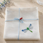 A folded white linen napkin with a blue dragonfly embroidery on the corner, tied with a blue string and a small red bow, set on a beige stone surface. MI RE LA