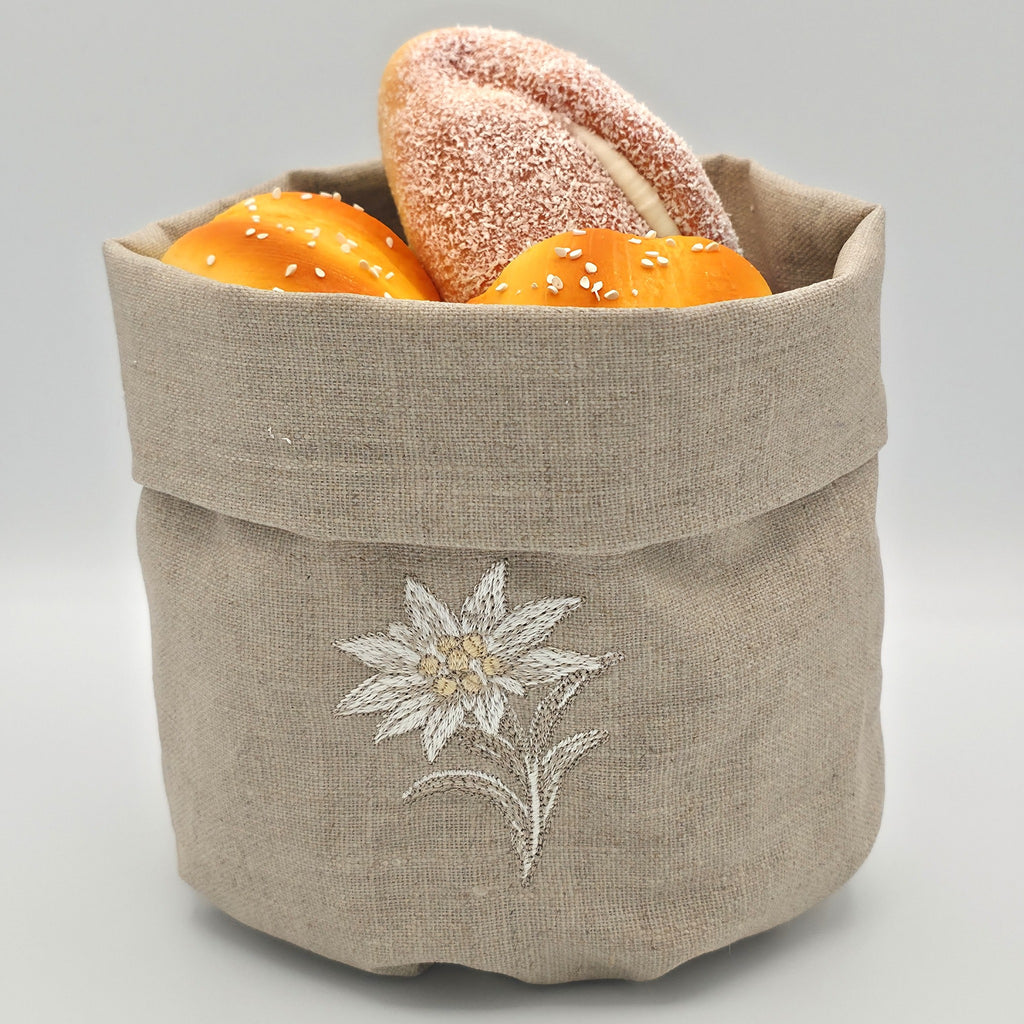 A oatmeal stone  linen bread basket with a white embroidered stella alpina on the front, filled with two bagels and two orange loaves, set against a neutral gray background.