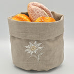 A oatmeal stone  linen bread basket with a white embroidered stella alpina on the front, filled with two bagels and two orange loaves, set against a neutral gray background.