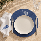 A styled dining place setting on a beige stone surface, featuring a navy blue round placemat with white jellyfish embroidery, a white ceramic plate, a white napkin with blue jellyfish embroidery, silver fork and knife, and a clear glass tumbler. MI RE LA