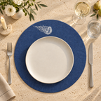 A styled table setting featuring a navy blue round placemat with a white jellyfish illustration, layered with a white ceramic plate, silver flatware, and dining glassware on a textured beige tablecloth.