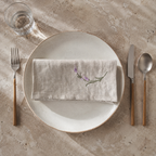 A styled flat-lay of a beige linen napkin with a small embroidered purple and green floral motif, folded on a round off-white ceramic plate. The setting includes a fork with a wooden handle on the left, a knife and a spoon with matching wooden handles on the right, and a glass of water on a textured beige stone surface. MI RE LA
