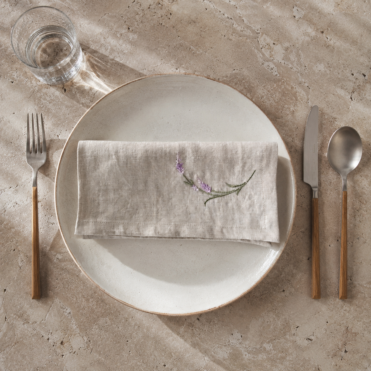 A styled flat-lay of a beige linen napkin with a small embroidered purple and green floral motif, folded on a round off-white ceramic plate. The setting includes a fork with a wooden handle on the left, a knife and a spoon with matching wooden handles on the right, and a glass of water on a textured beige stone surface. MI RE LA