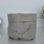 A light beige linen storage basket with a folded top, featuring an embroidered lavender branch design in purple and green on the front, set against a neutral light gray background.