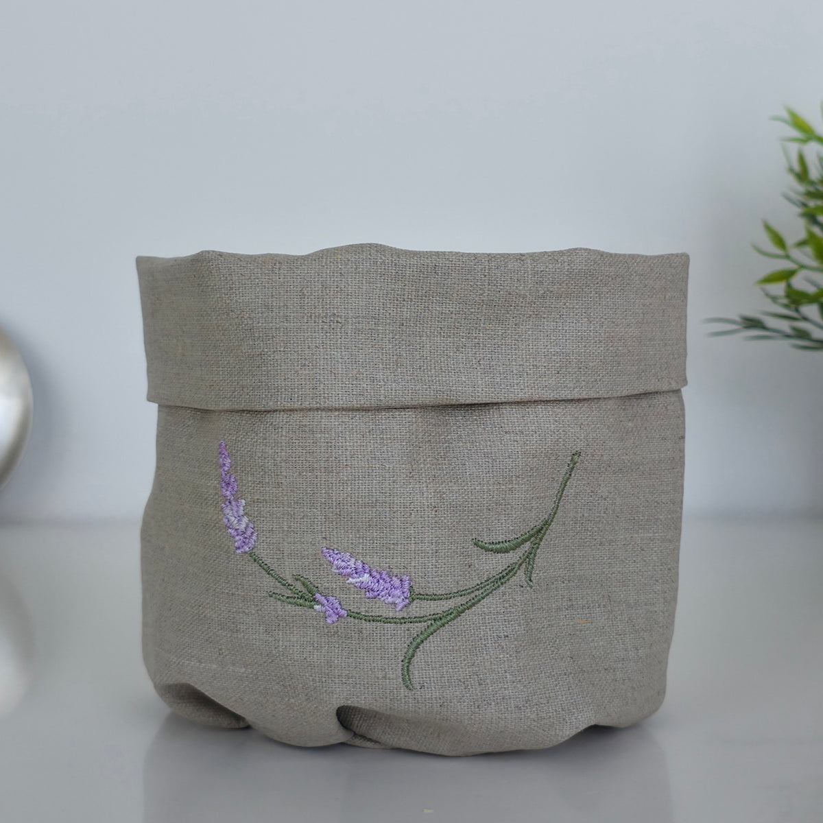 A light beige linen storage basket with a folded top, featuring an embroidered lavender branch design in purple and green on the front, set against a neutral light gray background.