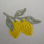 Close view of the lemon embroidery: two tapered yellow lemons on a branch with three sage green leaves, on off-white linen