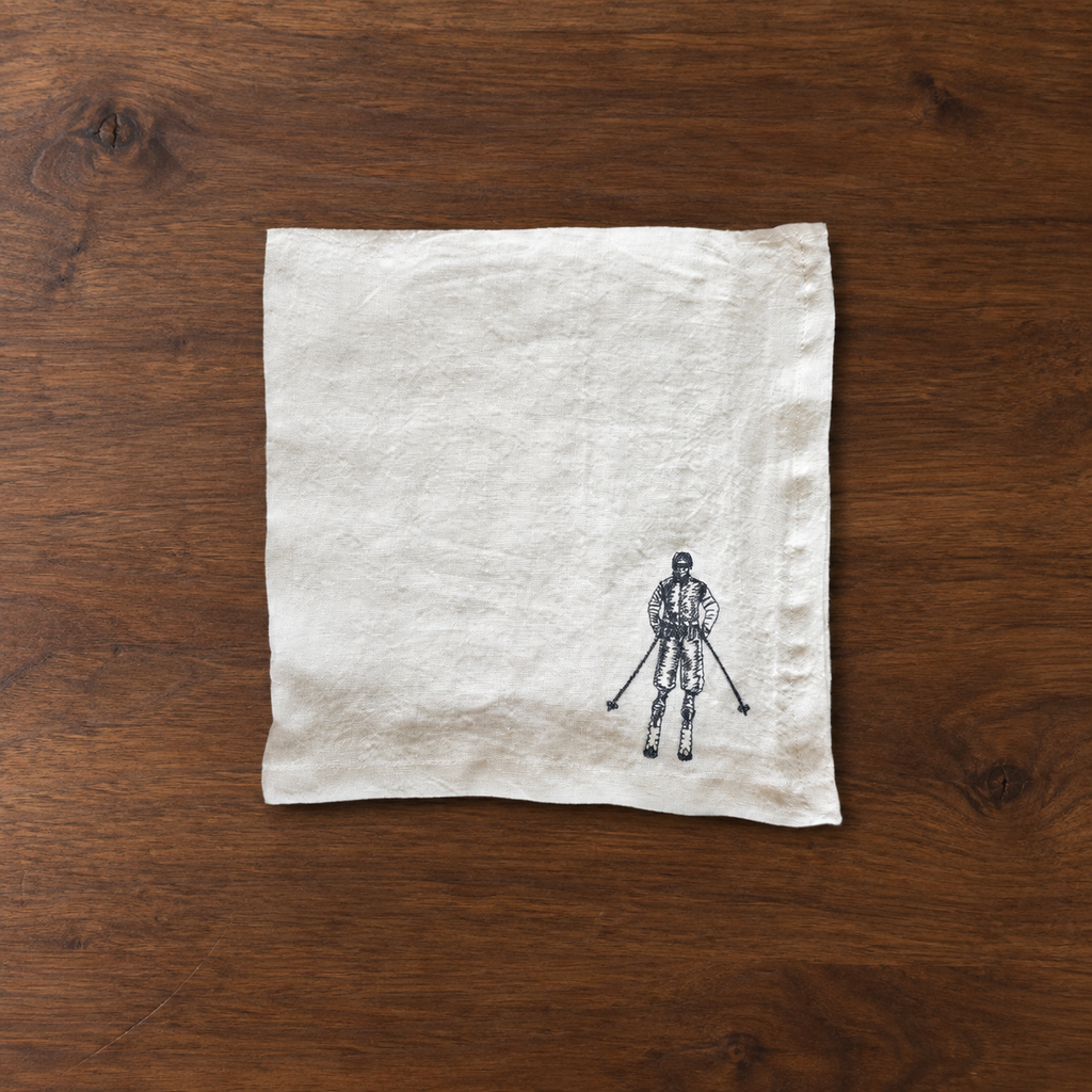 A square off-white linen napkin with a small black ink illustration of a skier at the bottom right, laid flat on a dark brown wooden surface. MI RE LA