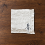 A square off-white linen napkin with a small black ink illustration of a skier at the bottom right, laid flat on a dark brown wooden surface. MI RE LA