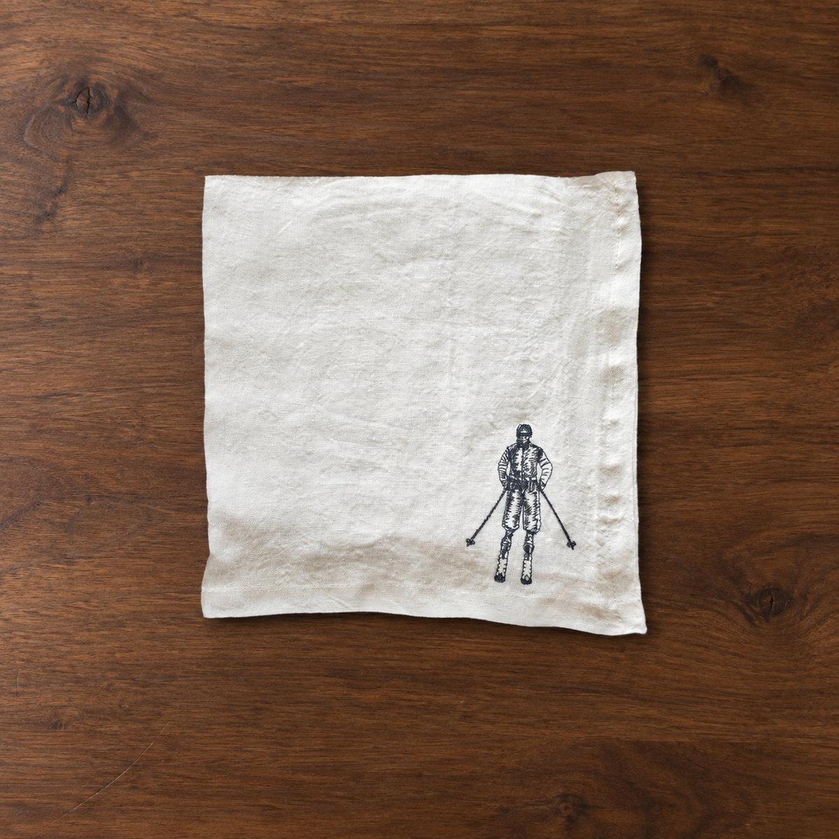 A square off-white linen napkin with a small black ink illustration of a skier at the bottom right, laid flat on a dark brown wooden surface. MI RE LA