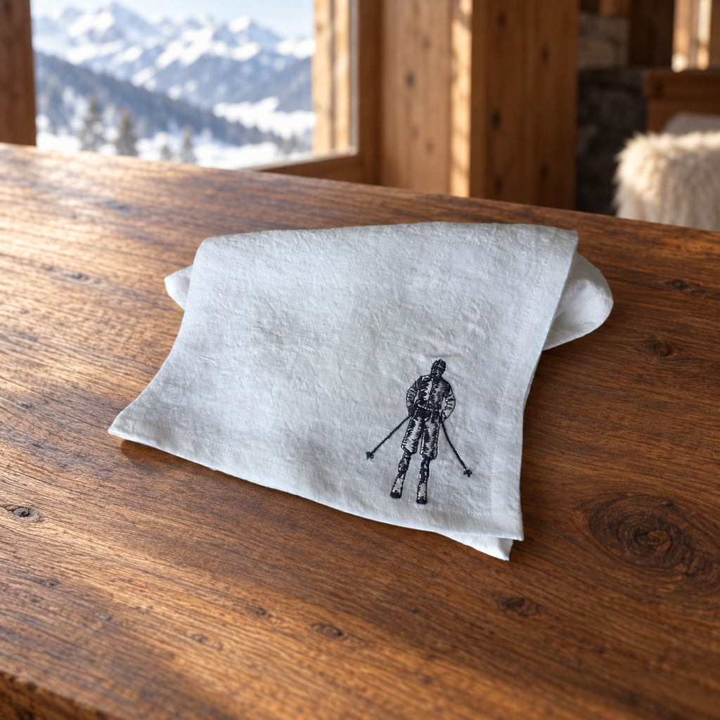 A light beige linen napkin with a black skier graphic, folded on a rustic wooden table with a snowy mountain window in the background. MI RE LA