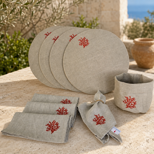 A linen tableware set displayed outdoors on a stone surface, including round plates, folded napkins, and a small cup or bowl, all in a light warm gray-beige fabric with small red coral embroidery.