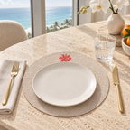 A place setting on a round beige dining table with a white plate on a woven beige placemat, a small red coral motif near the top, a gold fork on a folded cream napkin, a gold knife, a glass of water, and a vase with white orchids. A large window behind shows the ocean and palm trees.