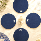 Four navy blue round coasters with white embroidered crab motifs, arranged in a diamond pattern on a light beige textured surface with decorative seashell and plant elements.