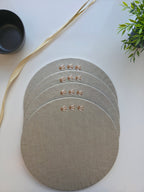 Four round linen placemats in a light taupe-beige color, stacked slightly offset on a white surface. Each placemat features a small embroidered gold monogram logo. MI RE LA