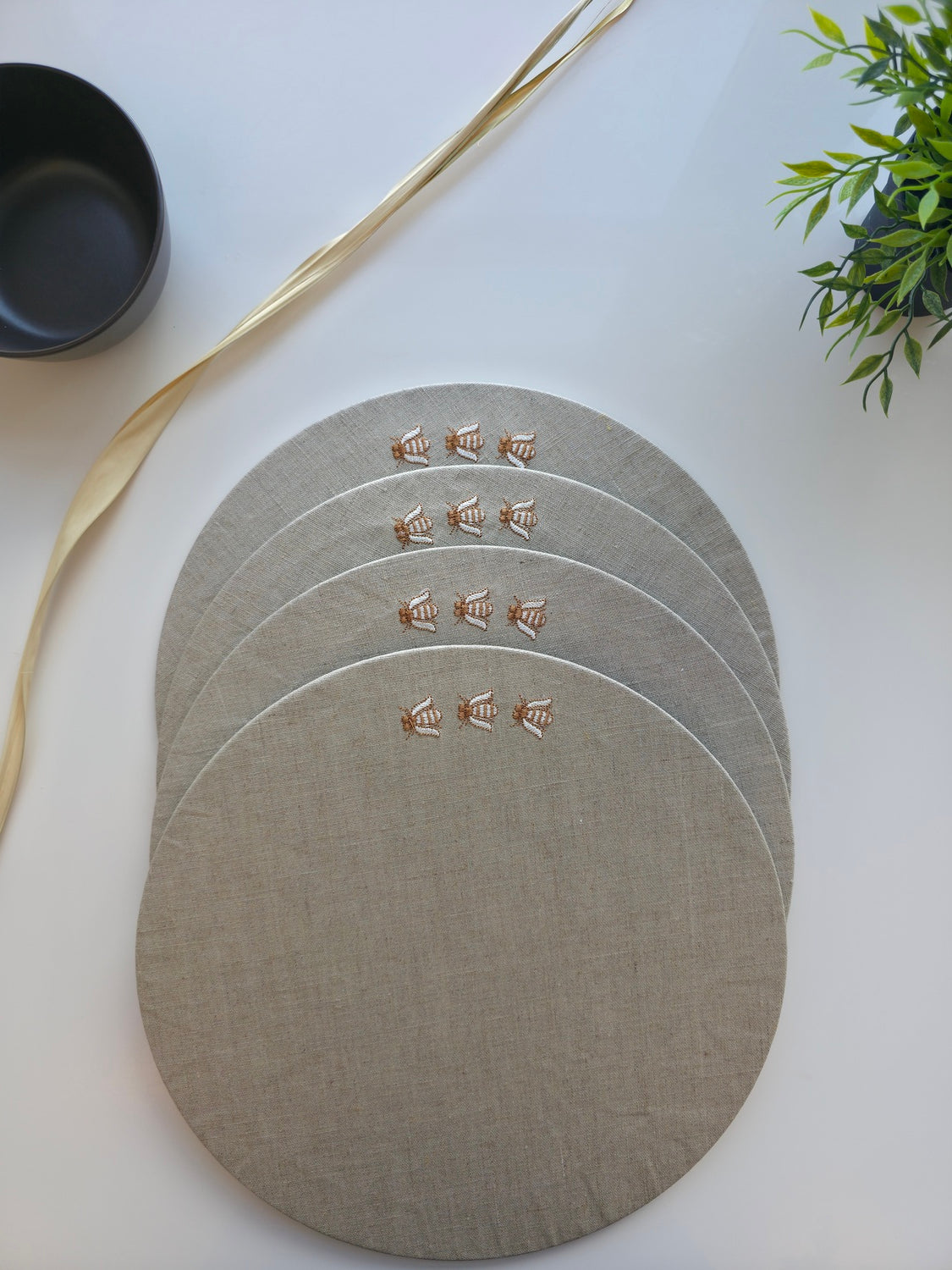 Four round linen placemats in a light taupe-beige color, stacked slightly offset on a white surface. Each placemat features a small embroidered gold monogram logo. MI RE LA