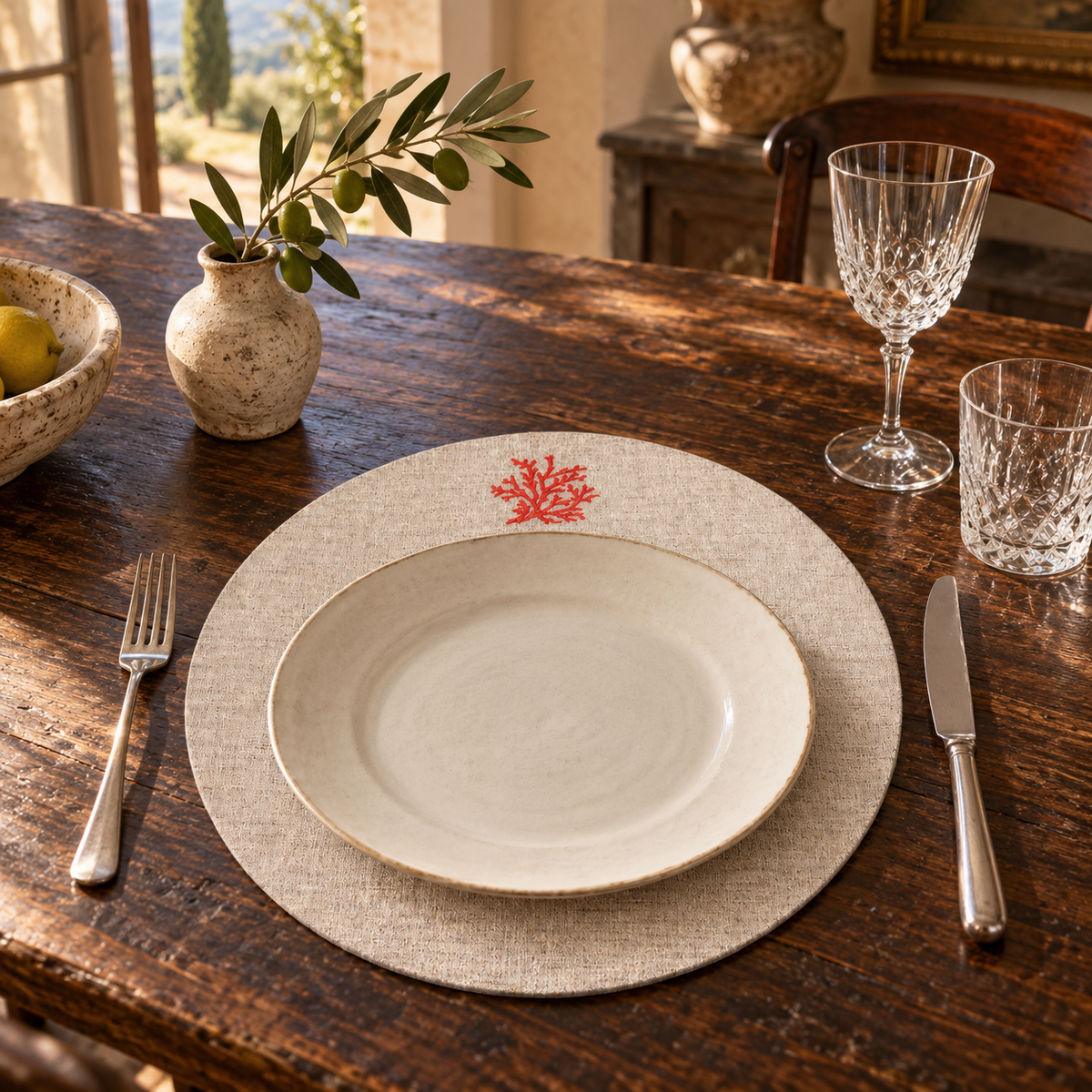 A place setting on a rustic dark wood dining table, featuring a round beige linen placemat with a small red coral motif, a cream ceramic plate, a silver fork, and a silver knife, with crystal glasses and a small vase in the background.