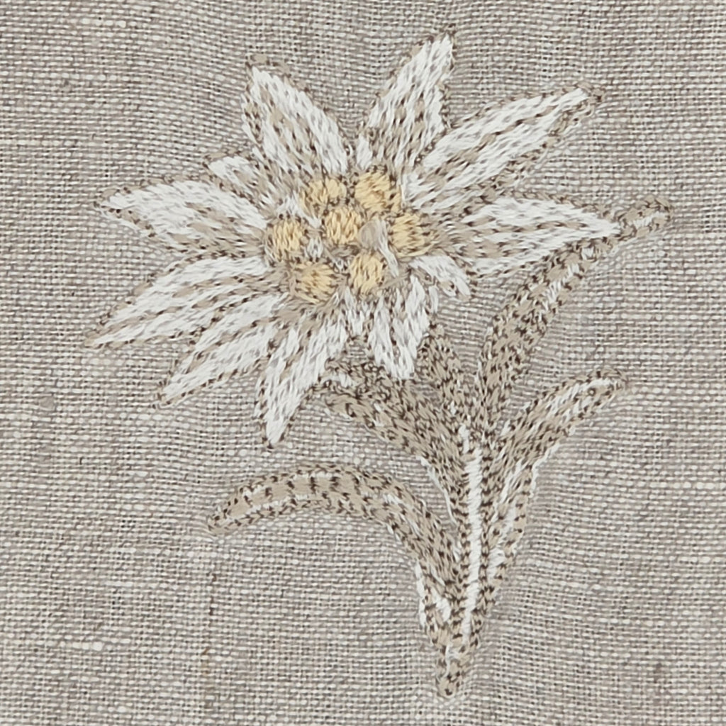Close-up of a edelweiss stella alpinaembroidery on a textured beige linen fabric, featuring cream and light yellow petals with brown and beige leaves. MI RE LA
