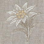 Close-up of a edelweiss stella alpinaembroidery on a textured beige linen fabric, featuring cream and light yellow petals with brown and beige leaves. MI RE LA