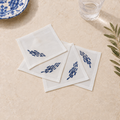 Four white square cotton napkins with blue embroidered fish motifs, arranged on a beige stone surface with a glass of water, a blue-and-white plate edge, and green leaves as props.