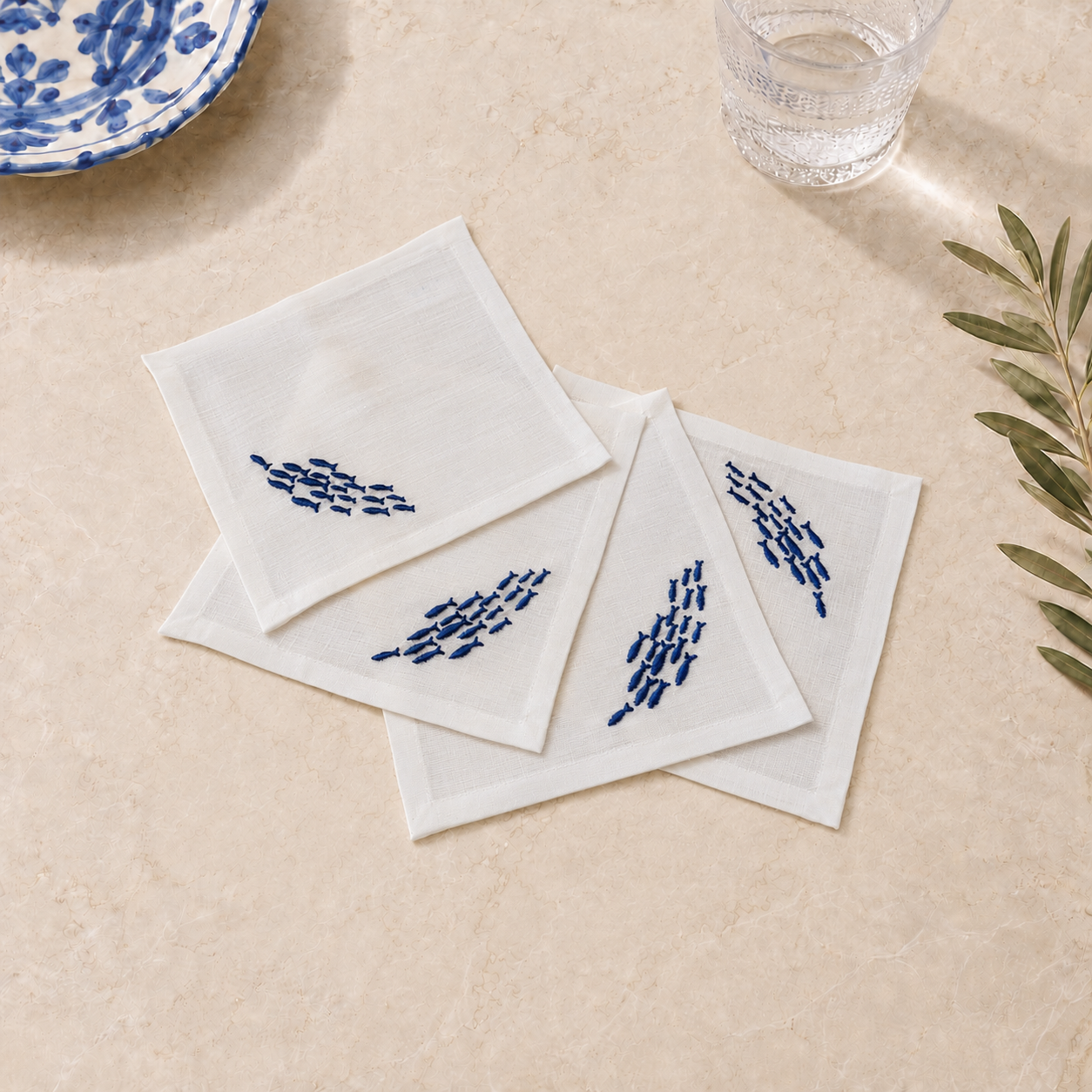 Four white square cotton napkins with blue embroidered fish motifs, arranged on a beige stone surface with a glass of water, a blue-and-white plate edge, and green leaves as props.