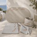 A coordinated linen table setting displayed outdoors against a textured cream wall, with a sea and distant land visible in the background. The set includes round placemats, folded napkins, and a small fabric basket, all in a light warm neutral tone with small embroidered floral details.