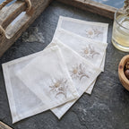 Three square off-white linen napkins with embroidered daisy-like floral motifs, arranged on a dark stone surface with rustic wooden props and a glass of pale liquid nearby.