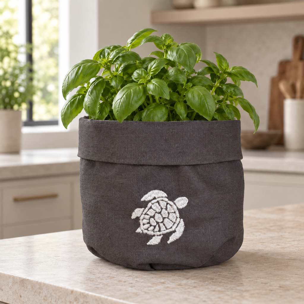 A dark gray fabric plant pot with a white embroidered sea turtle design, filled with a green basil plant and sitting on a kitchen countertop. MI RE LA
