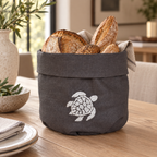 A dark gray fabric bread basket with a white turtle design, filled with crusty bread loaves and set on a wooden dining table in a warm, modern kitchen setting. MI RE LA