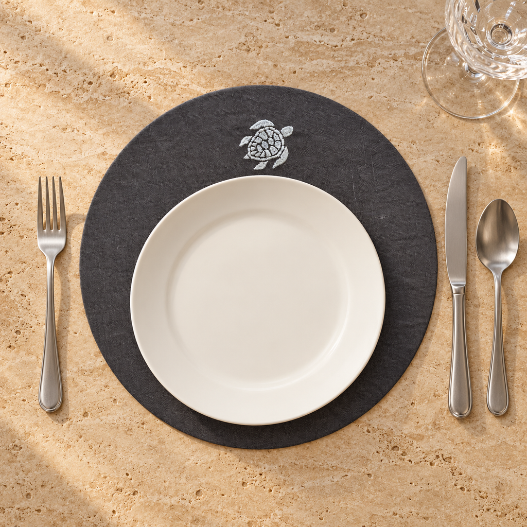 A place setting on a beige stone surface featuring a round black placemat with a white sea turtle emblem, a white ceramic plate, silver flatware, and a clear wine glass. MI RE LA