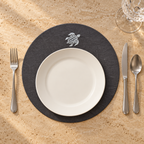 A place setting on a beige stone surface featuring a round black placemat with a white sea turtle emblem, a white ceramic plate, silver flatware, and a clear wine glass. MI RE LA
