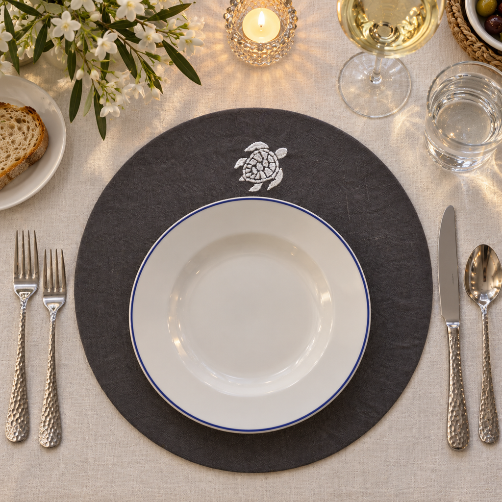 A styled table setting featuring a round dark gray placemat with a white sea turtle emblem, layered with a white ceramic plate that has a thin blue rim. The setting includes silver cutlery, a glass of white wine, a candle, and a linen tablecloth. MI RE LA