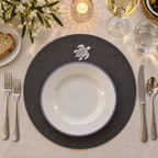A styled table setting featuring a round dark gray placemat with a white sea turtle emblem, layered with a white ceramic plate that has a thin blue rim. The setting includes silver cutlery, a glass of white wine, a candle, and a linen tablecloth. MI RE LA