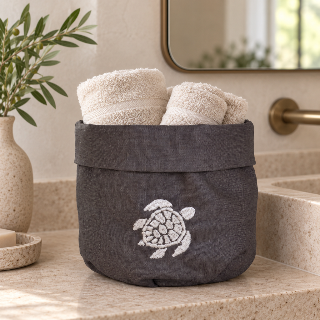 A dark gray fabric bath towel basket with a white embroidered sea turtle, filled with cream-colored towels and sitting on a beige stone bathroom counter. MI RE LA